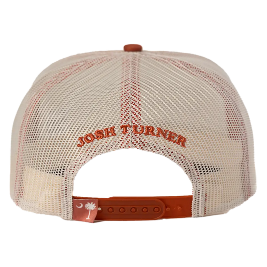 Cream-colored mesh trucker hat with orange embroidered ’josh turner’ text and a matching orange adjustable strap featuring a white crescent moon and palmetto tree logo.