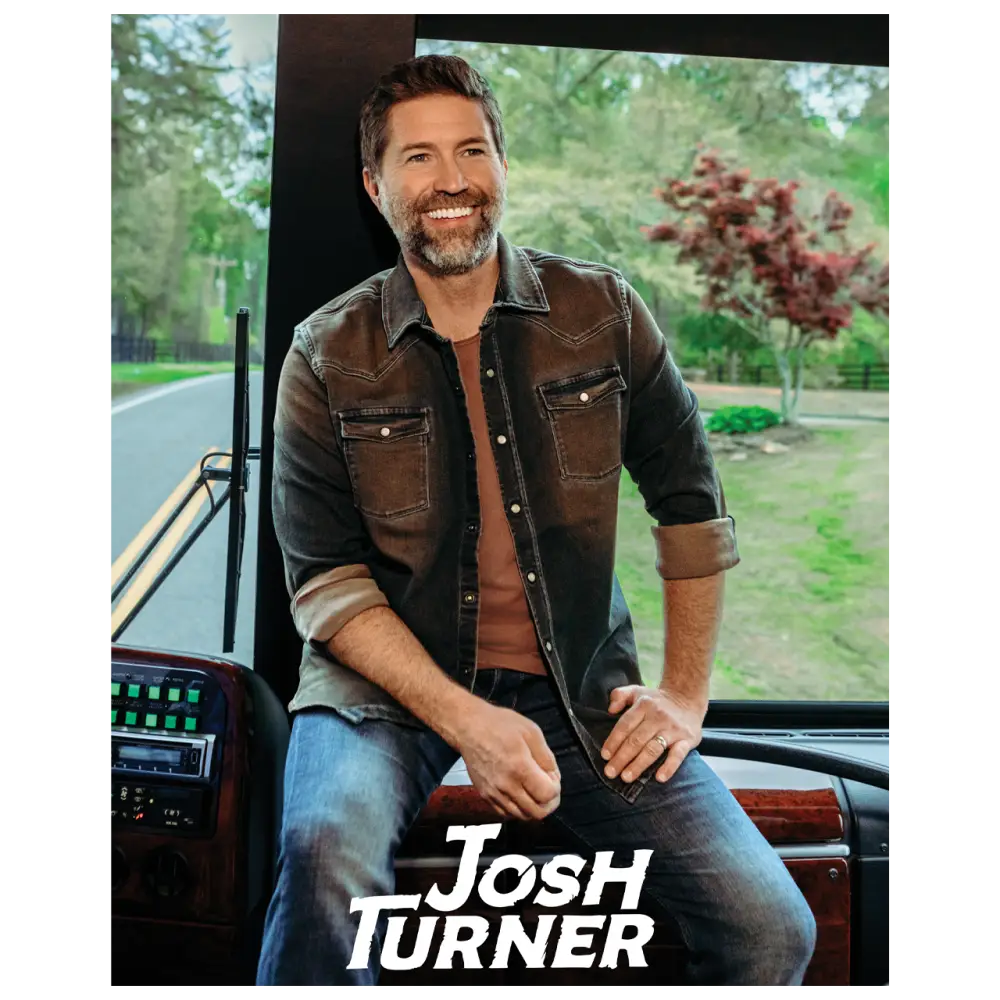 Smiling man with a beard wearing a brown jacket and jeans seated in a vehicle.