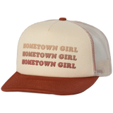 Cream-colored trucker hat with a rust-brown brim and mesh back, featuring the phrase ’hometown girl’ embroidered in three stacked rows of matching rust-brown lettering.