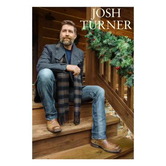 Josh turner sits on wooden steps wearing a dark gray woolen jacket, blue jeans, and brown leather boots with a plaid scarf draped over his lap.