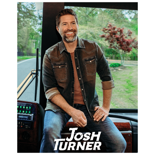 Smiling man with a beard wearing a brown jacket and jeans seated in a vehicle.