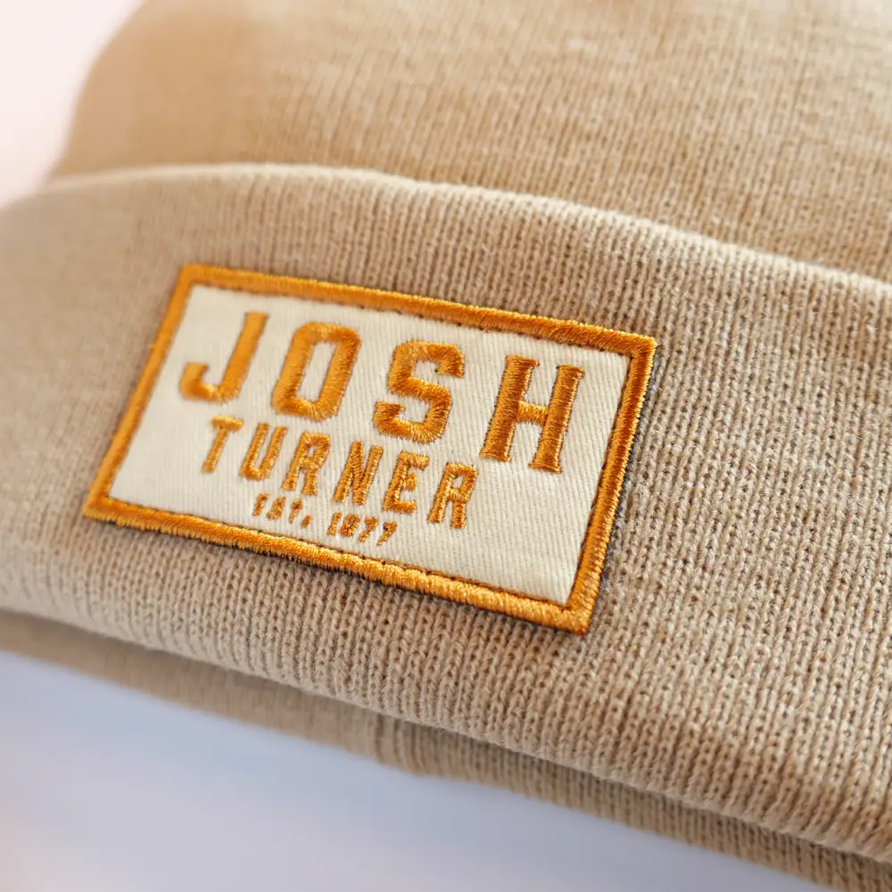 Beige knit beanie with an orange and white embroidered patch reading ’JOSH TURNER EST. 1977’’.