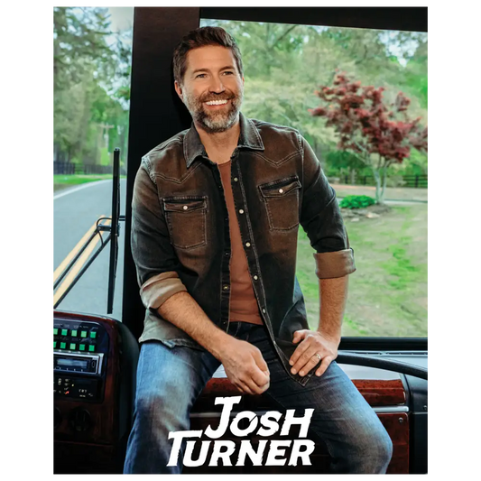 Smiling man with a beard wearing a brown jacket and jeans seated in a vehicle.