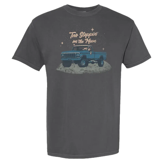 Dark gray t-shirt featuring a cartoon illustration of a blue pickup truck on the moon with the text ’two steppin’ on the moon’ above it.